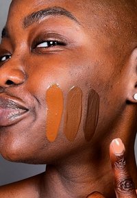 Fyra grundfärgsprov i olika bruna nyanser applicerade på en kind, som visar en krämig konsistens. Slät hud och en handgest ingår.