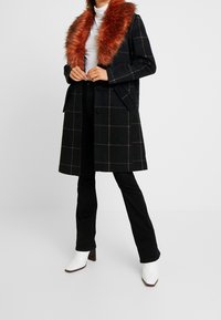 Manteau long à carreaux noirs et marrons avec un large col en fausse fourrure couleur rouille, porté sur un col roulé blanc, associé à un pantalon noir et des bottes blanches.