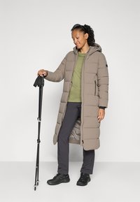 Lange, bruine pufferjas met een capuchon, ritssluiting en twee zijzakken, gedragen over een groene top en zwarte broek, met trekkingstokken in de hand.