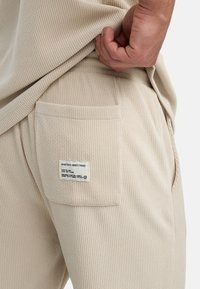 Beige strukturierte Jogginghosen mit einer sichtbaren Gesäßtasche, die ein Etikett besitzt; das Material zeigt ein dezentes Rippenmuster. Entworfen für Komfort und Langlebigkeit.