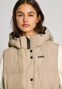 Beige vadderad väst med hög krage och avtagbar huva, med svarta knappar och dragkedja, samt ett litet logopatch. Texturerad tyg.