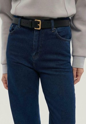 Cintura in pelle nera con fibbia in metallo dorato, indossata sopra jeans blu scuro, caratterizzati da un taglio classico dritto e tasche laterali.