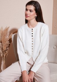 Femme aux longs cheveux foncés portant un cardigan blanc boutonné et un pantalon beige clair, assise sur une chaise beige avec des plantes sèches à proximité.