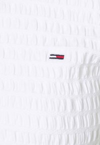 Vit texturerad tyg med litet broderat rektangulärt logotyp som har svarta, vita och röda horisontella ränder.