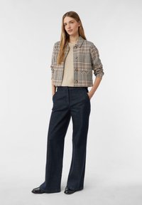 Žena stojící s rukama v kapsách, oblečená v béžovém svetru, kostkovaném zapínacím saku, tmavých širokých kalhotách a černých botách na bílém pozadí.