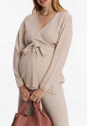 Beige wrap sweater with long sleeves, tied at waist, made of soft material. A matching bottom and a pink handbag are held in hands.