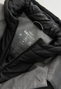 Interior acolchado negro con tela texturizada gris. Presenta una etiqueta de marca "NEXT DRYTECH", cremallera y lazo de cordón. Indica tamaño M.