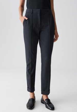 Vrouw in donkergrijze, op maat gemaakte, omgeslagen broek, met zwarte instappers en een zwart mouwloos topje, staand met één hand in de zak.