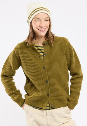 Lachende vrouw met een crèmekleurige gebreide muts, een olijfgroen vest met knopen over een groen gestreept overhemd en beige broek met handen in de zakken.