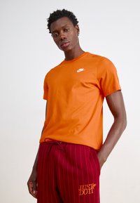 Orange cotton t-shirt with a round neckline and short sleeves, featuring a white Nike logo on the chest, paired with red striped pants.