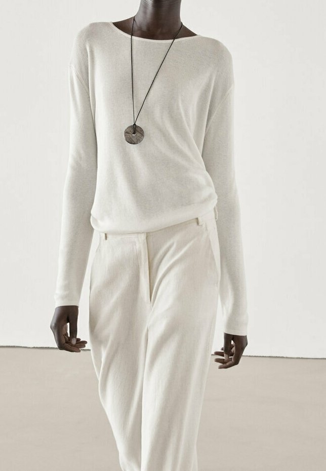 Personne portant un pull crème à manches longues et un pantalon assorti, accessoirisé d'un collier pendentif rond sur un cordon noir.