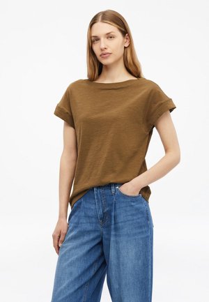 Kobieta z prostymi, jasno brązowymi włosami ma na sobie luźny brązowy T-shirt i niebieskie dżinsy, stoi na tle gładkiej białej ściany, ręka w kieszeni.