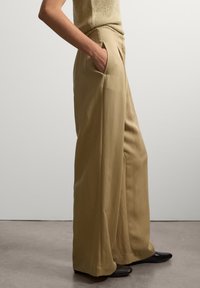 Weite Hosen aus khakifarbenem Stoff, mit Seitentaschen und glatter Textur. Kombiniert mit spitzen schwarzen Schuhen. Minimalistisches Design.