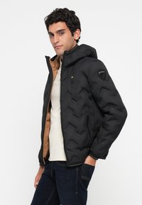 Sort jakke af puffer-typen med hætte, i en chevron-mønster, to-farvet design med indvendig brun foring og lynlåslukning.
