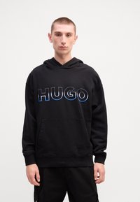 Svart hettegenser med en brystlomme; har fet blå og hvit "HUGO" tekst over brystet; mykt, strukturert stoff.