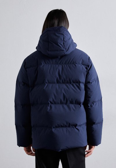 Doudoune bleu marine avec capuche, présentant des sections matelassées et une surface lisse et texturée. Design simple avec une coupe décontractée.