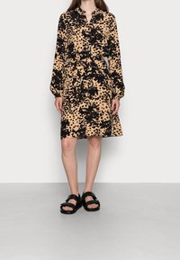 Beige skjortklänning med långa ärmar och svart leopardmönster, har en knappslå, bälte i midjan och smockade ärmar, kombinerad med svarta sandaler.