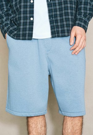 Man wearing light blue knee-length shorts with hands in pockets, paired with a white shirt and dark green plaid button-up shirt.