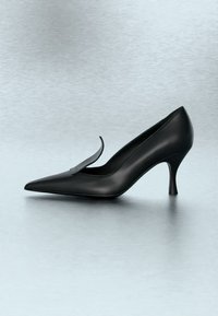 Chaussure à talon haut noire pointue avec un détail de bride courbée, présentée sur une surface lisse gris clair.