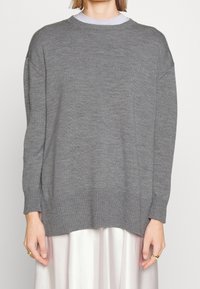 Eine Frau trägt einen lockeren grauen Strickpulli über einem weißen Rock, mit goldenen Ringen an beiden Händen vor einem einfarbigen Hintergrund.