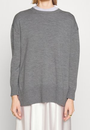 Vrouw in een losse grijze gebreide trui over een witte rok, met gouden ringen aan beide handen tegen een effen achtergrond.