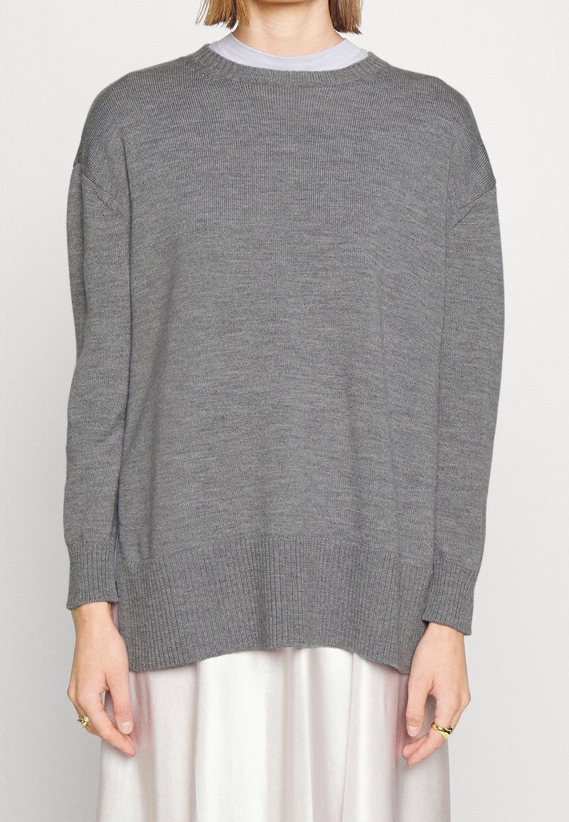 Eine Frau trägt einen lockeren grauen Strickpulli über einem weißen Rock, mit goldenen Ringen an beiden Händen vor einem einfarbigen Hintergrund.