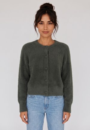 Donna con capelli scuri raccolti in uno chignon che indossa un maglione verde scuro morbido e con bottoni e jeans azzurri, rivolta in avanti su uno sfondo semplice.
