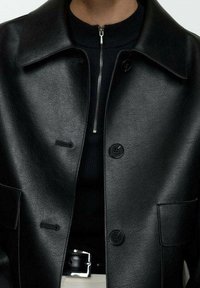 Sort læderjakke med krave, frontknaplukning, sidelommer og en glat struktur. Lagt over en mørk top med lynlås i halsen.