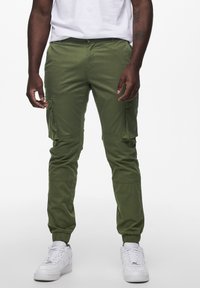 Groene cargo-broek met elastische boorden, voorzien van meerdere zijzakken en een gladde katoenen stof. Gepaarde met witte sneakers.
