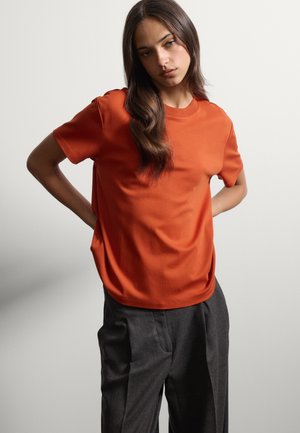 Jonge vrouw met lang donker haar, gekleed in een oranje T-shirt met korte mouwen en donkergrijze broek, staand met haar handen achter haar rug.