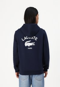 Tamsiai mėlynas gobtuvėlis, pagamintas iš minkštos medžiagos. Ant nugaros yra baltas tekstas "LACOSTE PARIS" ir krokodilo logotipas. Klasikinis dizainas ir tekstūruotas paviršius.