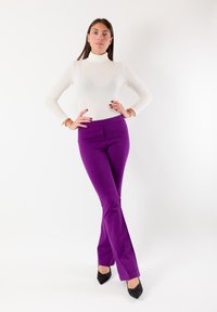 Maglione dolcevita bianco, pantaloni svasati viola, vestibilità slim. Scarpe nere piatte con punta appuntita. Modella in piedi con le mani sui fianchi su sfondo bianco.