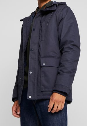 Homme portant une veste d'hiver à capuche bleu marine foncé avec fermeture à boutons et fermeture éclair, poche à rabat, et jean, sur un fond uni.