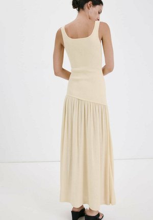 Woman seen from behind wearing sleeveless beige maxi dress with gathered skirt and black heeled sandals against plain white background.