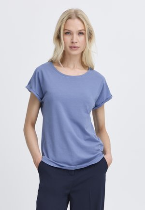 Blondýna s rozpuštěnými vlasy, oblečená v modrém tričku s krátkými rukávy a tmavých kalhotách, stojící s rukama v kapsách na šedém pozadí.