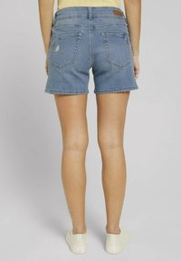 Donna che indossa shorts di denim blu chiaro con un piccolo strappo sul taschino posteriore sinistro e sneakers bianche, in piedi contro uno sfondo neutro.