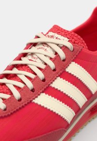 Zapatilla deportiva roja y rosa con una parte superior texturizada, tres franjas blancas y cordones beige. Suela de goma con un patrón de relieve distintivo.