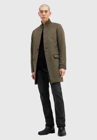 Cappotto in misto lana verde oliva con colletto alzato, chiusura con un solo bottone e due tasche frontali, abbinato a jeans neri e stivali neri.