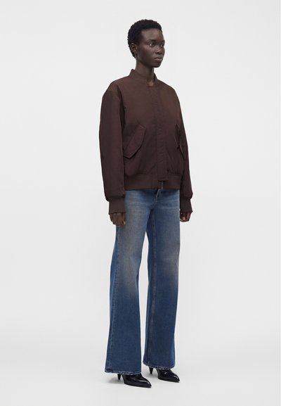 Γυναίκα φοράει σκούρο καφέ bomber μπουφάν, μπλε τζιν με ψηλή μέση και φαρδιά πόδια, και μαύρα ανδρικά παπούτσια με μυτερά ακροδάχτυλα, στέκεται μπροστά σε λευκό φόντο.