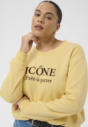 Frau mit zurückgebundenem Haar, die einen gelben Pullover mit dem Text „ICÔNE Prêt-à-porter“ trägt, eine Hand nahe am Kinn, die andere verschränkt.