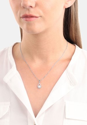 Silberne Halskette mit einem tropfenförmigen Anhänger, der eine zentrale Perle zeigt, umgeben von einer zarten Kette, getragen über einer weißen Bluse.