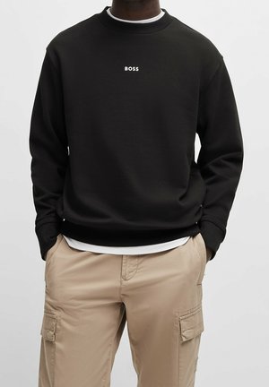 Homme portant un sweat-shirt noir avec le texte « BOSS » et un pantalon cargo beige, mains dans les poches, fond neutre.