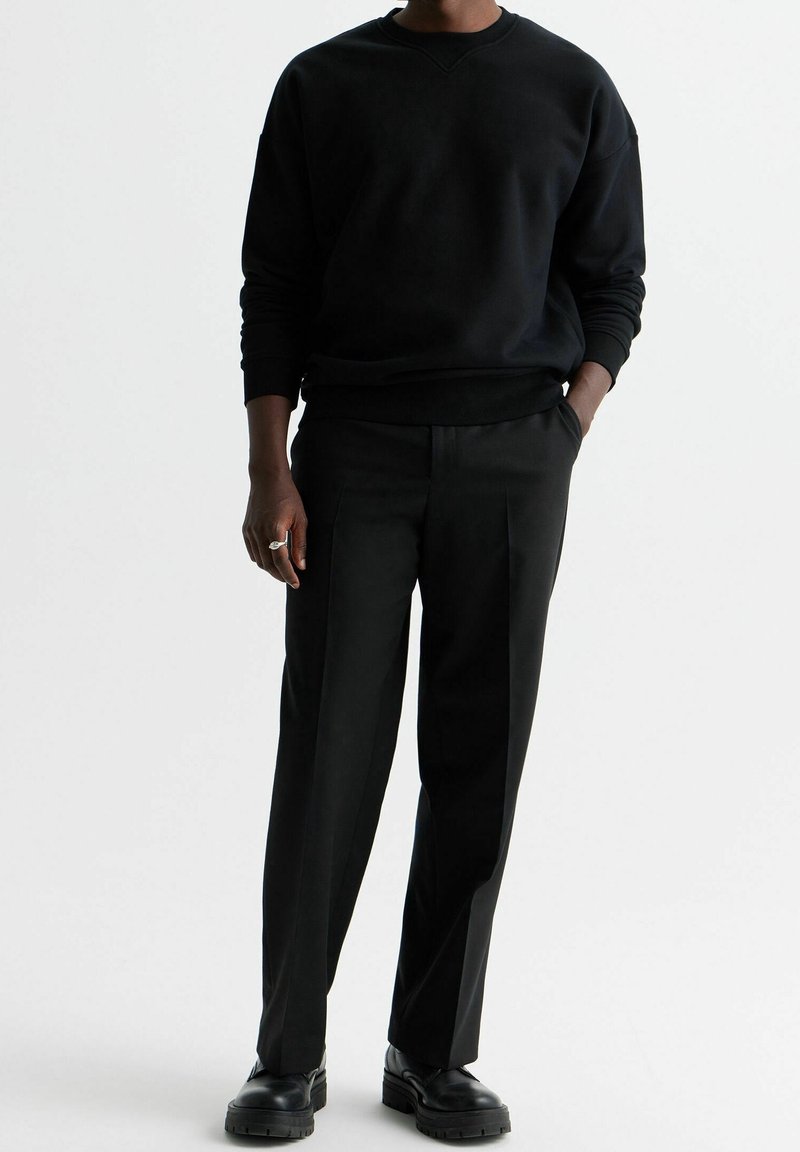 Personne portant un sweat-shirt noir à manches longues, un pantalon plissé noir, des chaussures en cuir noires, avec une main dans la poche et une main visible.