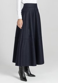 Kobieta ubrana w granatową spódnicę midi w kształcie litery A zestawioną z czarnymi botkami na obcasach i białą koszulą z długim rękawem.