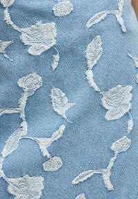 Tejido de mezclilla azul claro con patrones florales bordados en blanco. Textura desgastada con bordes deshilachados que resaltan hojas y brotes.