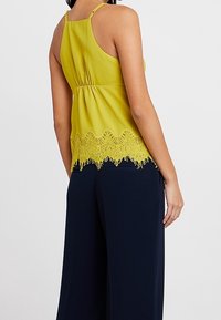 Haut sans manches jaune avec des embellissements en dentelle à l'ourlet, taille froncée et fines bretelles, associé à un pantalon large bleu marine.