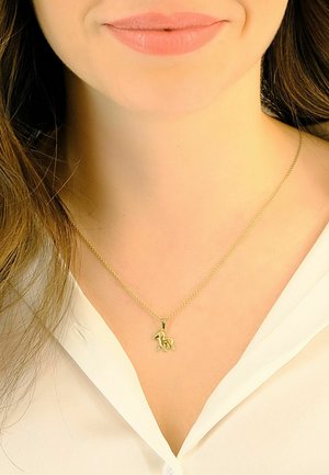 Collier pendentif ton or avec un charme en forme de cheval, suspendu à une chaîne délicate, porté sur une chemise blanche à col.