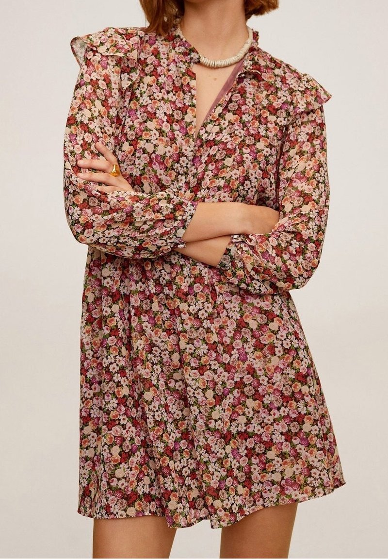 Femme portant une robe fleurie à manches longues avec des fleurs roses, rouges et blanches, bras croisés et un collier de coquillages blanc.