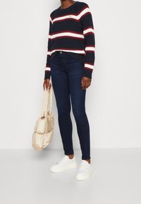 Persoon in een marineblauwe kabeltrui met strepen, donkere skinny jeans, witte platform sneakers, met een beige gevoerde draagtas aan de zij.