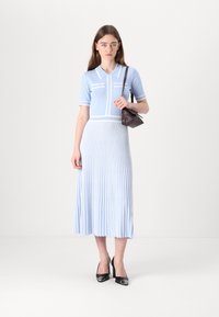 Ljusblå stickad klänning med korta ärmar, vita detaljer och vertikala veck; matchad med en liten mörk handväska och svarta klackar.